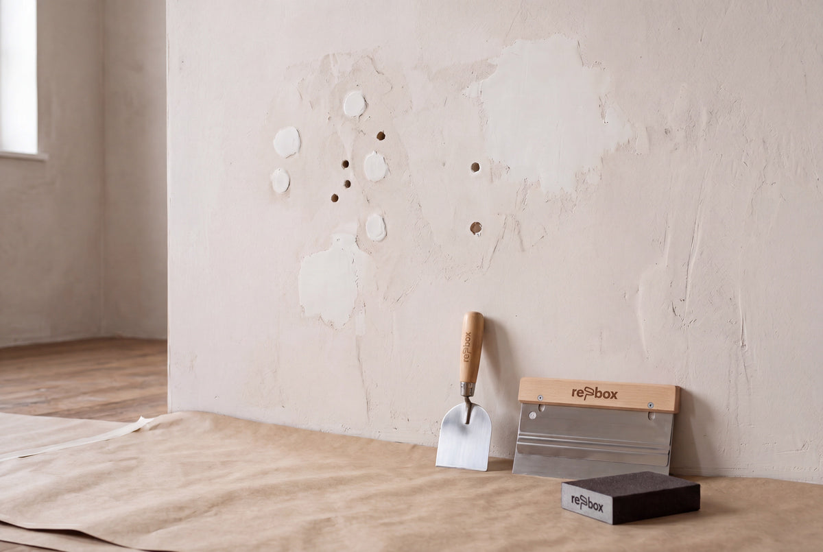 re-box – gespachtelte Wand mit gefüllten Bohrlöchern, dazu Glättekelle, Spachtel und Schleifschwamm